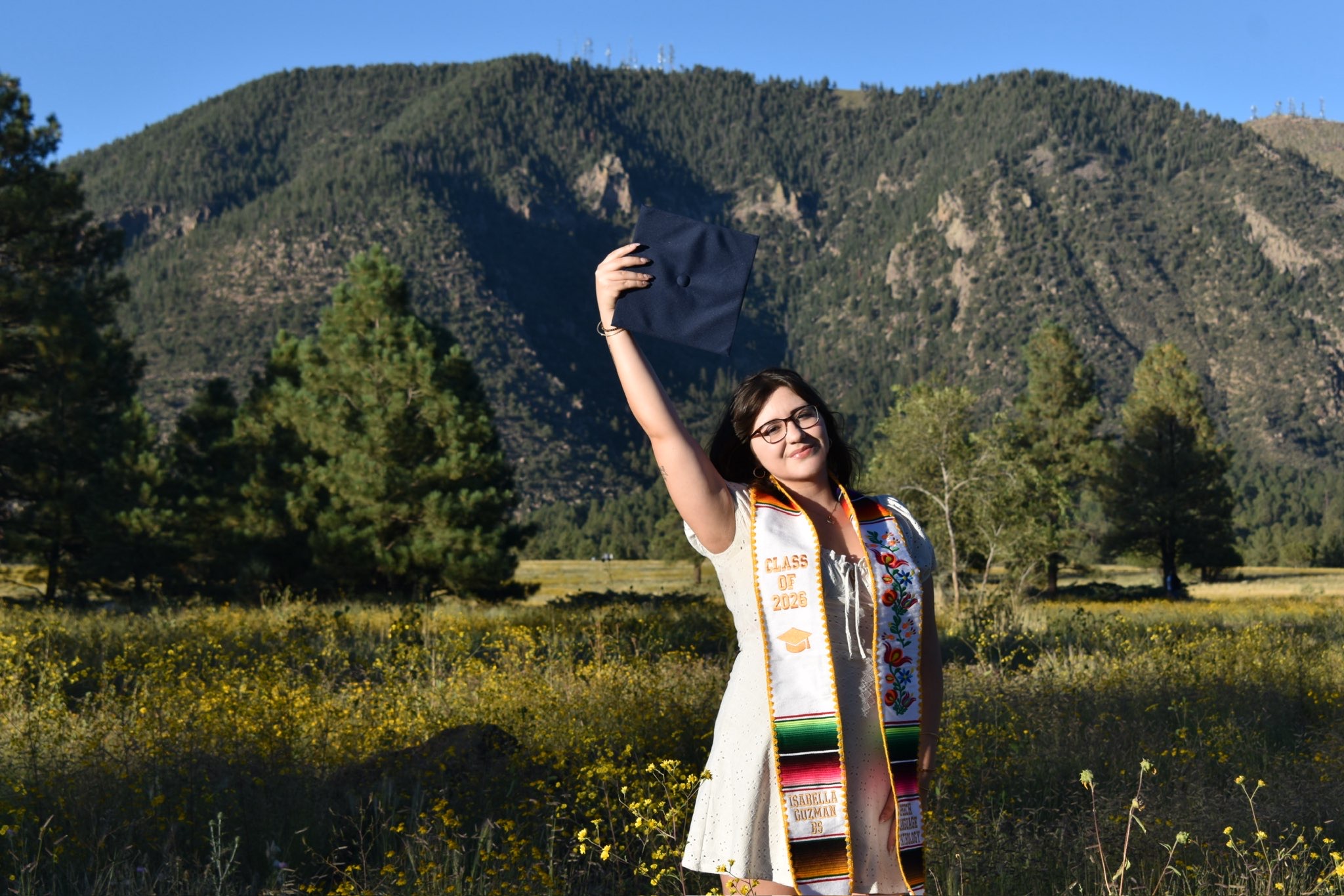 Isabella Guzman posing with graduation regalia in a meadow in front of Flagstaff mountains and blue sky.