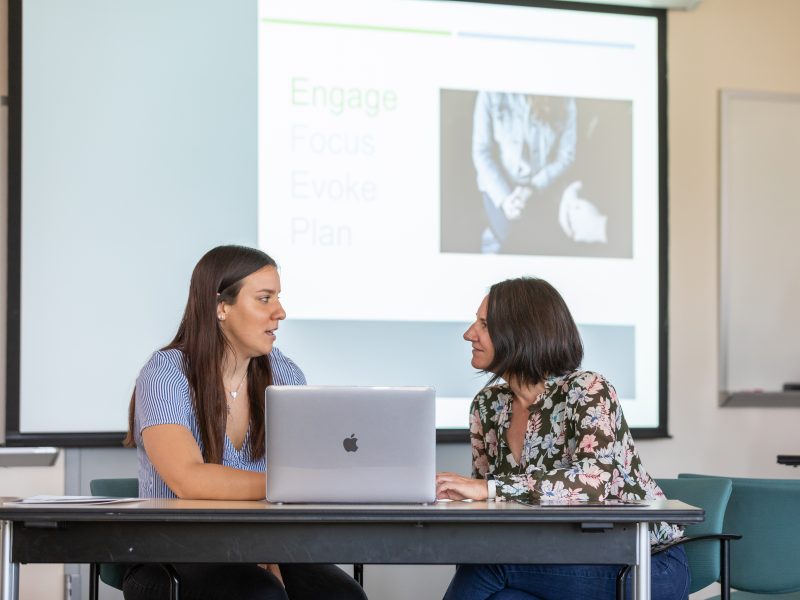 Faculty members speaking to each other in a classroom.