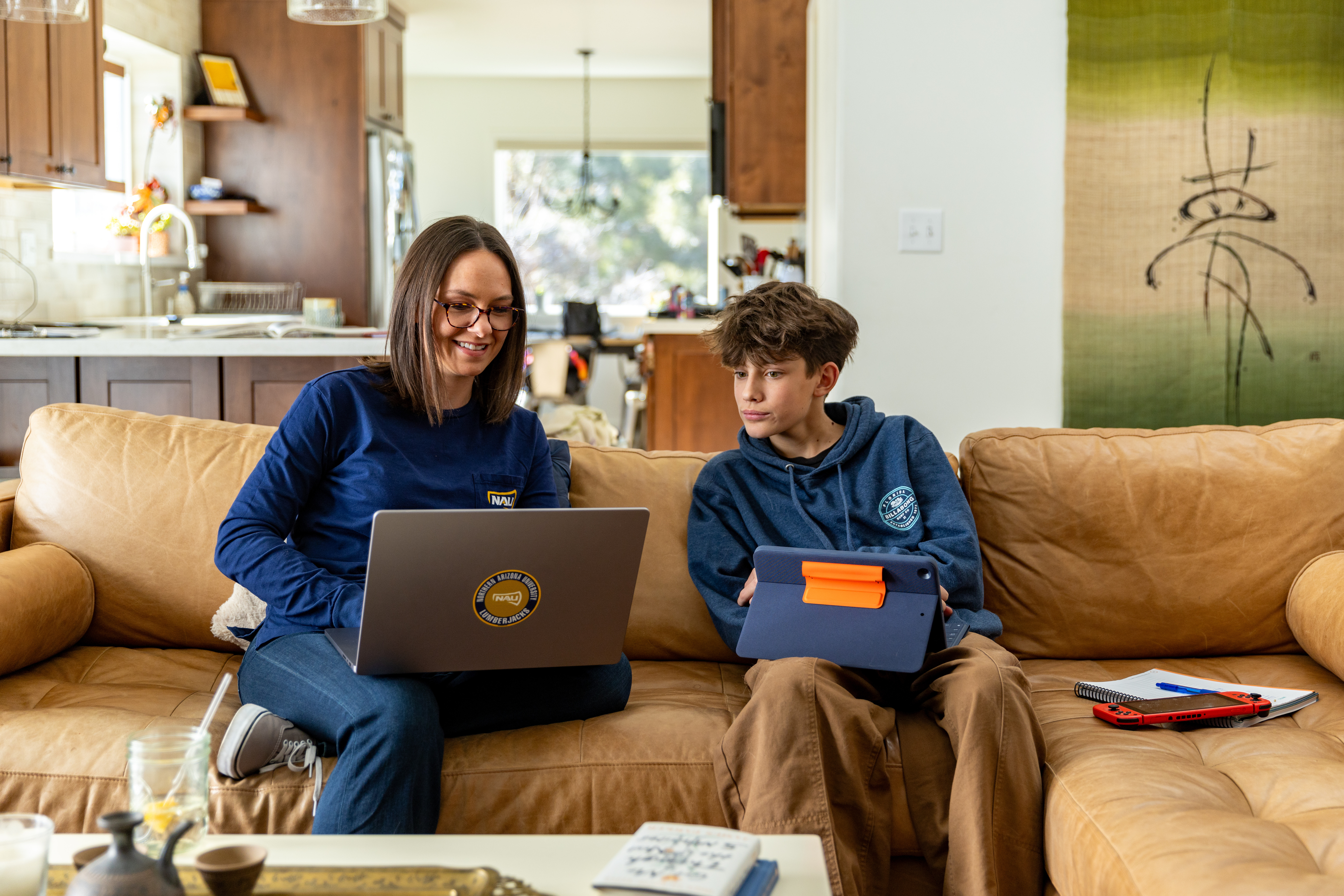 Two people sitting on a couch, using laptops. The person on the left is wearing a blue hooded sweatshirt and the person on the right is wearing a blue hooded sweatshirt. The room contains shelves, a refrigerator, and other household items.