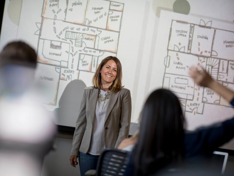 Faculty member standing in front of projector screens showing large interior design layouts.