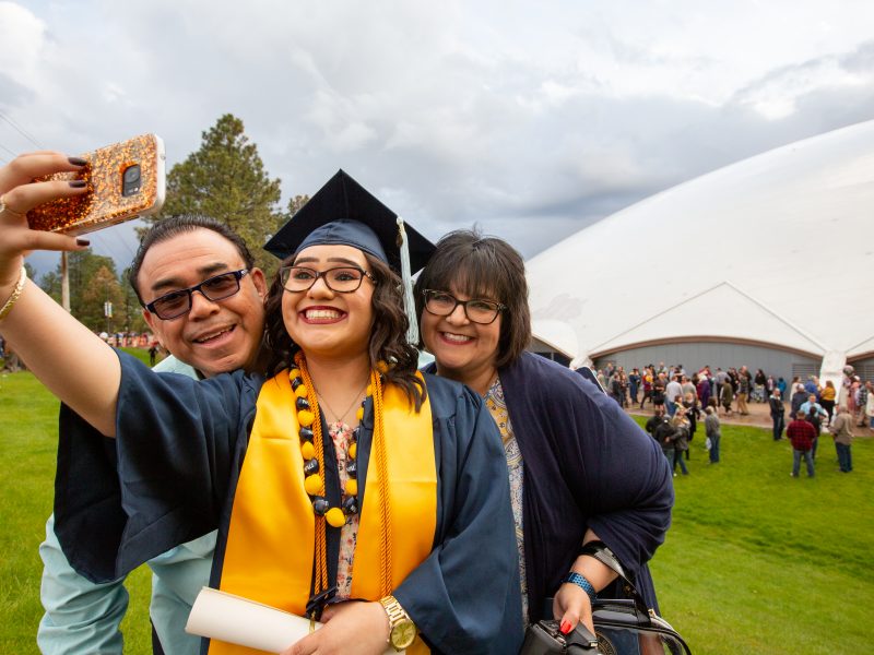 N A U graduate taking selfie in cap and gown outside commencement.