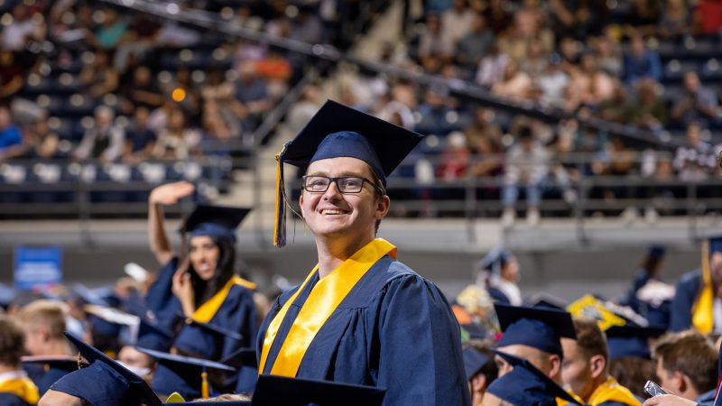 N A U graduate smiling in graduation regalia while other graduates sit waiting for the commencement ceremony to begin.