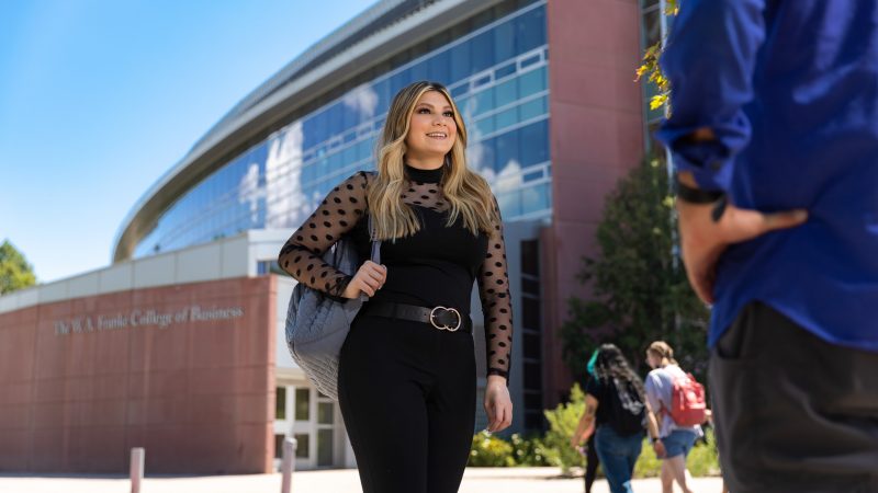 Smiling student walking in front of The W A Franke College of Business building.