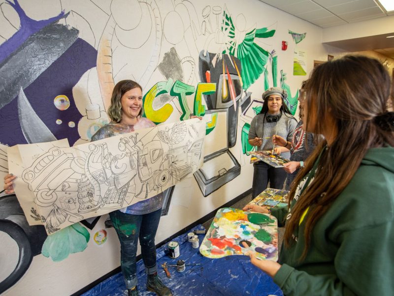 Students paint and display art on wall smiling.