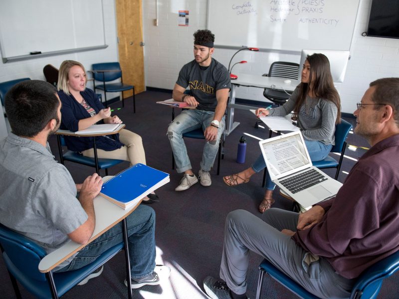 A group of students and professors sitting in desks in a circle talking to each other.