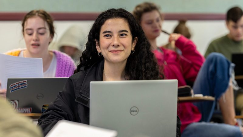 A person sitting at a classroom desk with a laptop smiling.