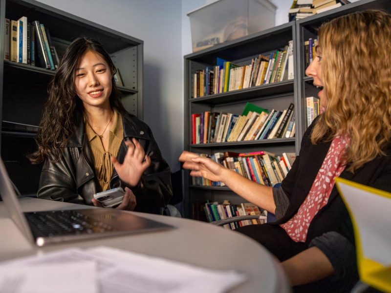 A person wearing a black leather jacket is sitting at a table with a professor looking at a laptop screen.