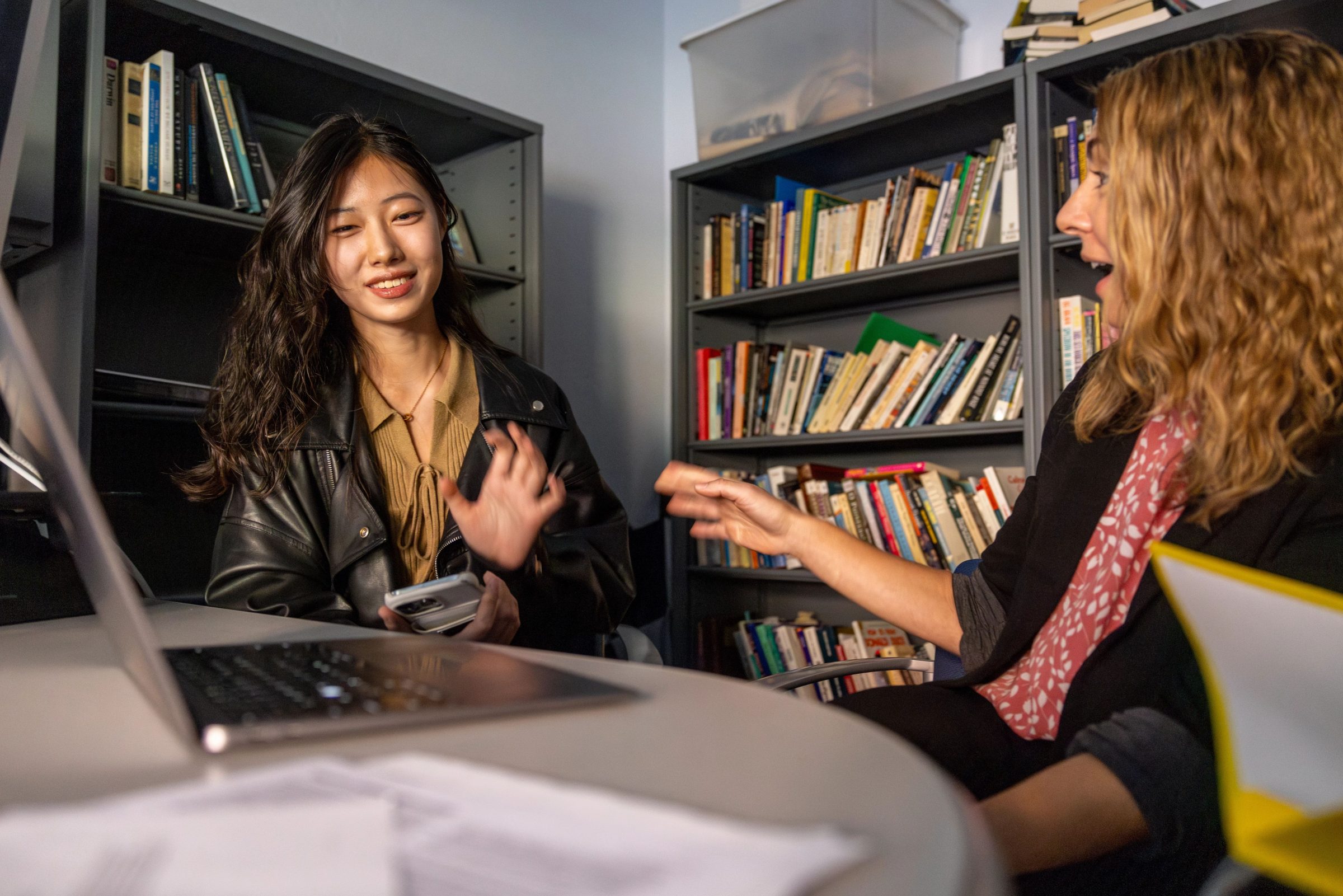 A person wearing a black leather jacket is sitting at a table with a professor looking at a laptop screen.