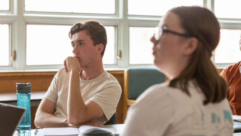 Students in white shirts are sitting at a desk, gazing towards a presentation.