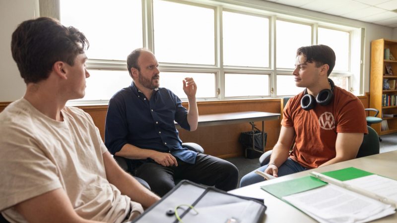 A person wearing headphones around their neck is sitting at a table talking to a professor in a blue shirt.