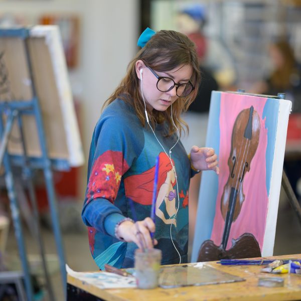 Student adding more paint to her brush while working on a painting of a violin.
