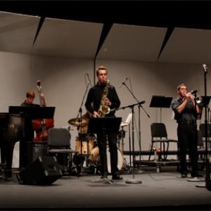 student plays saxophone on stage while student plays the cello behind him