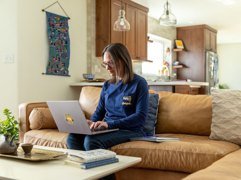 Student sitting on a couch typing on a laptop computer in a living room setting, with shelves, plants, and lighting fixtures visible.