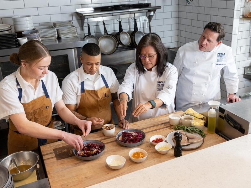 Four people in chef's uniforms working together in a kitchen, preparing food on a wooden counter. Pots, pans, and utensils are visible on the shelves behind them. The counter contains various ingredients and dishes.