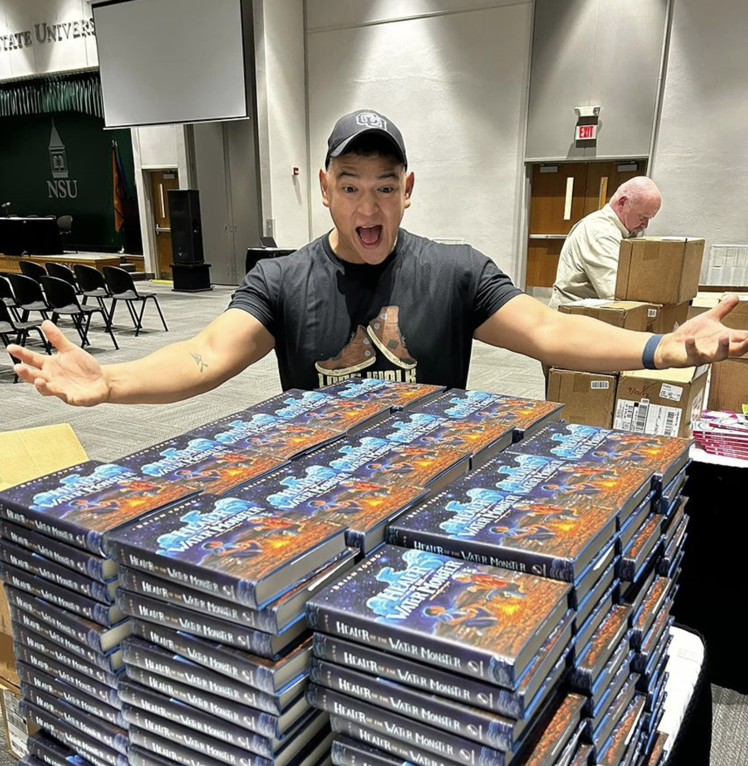 Brian Young behind stacks of his book at a speaking engagement looking excited.