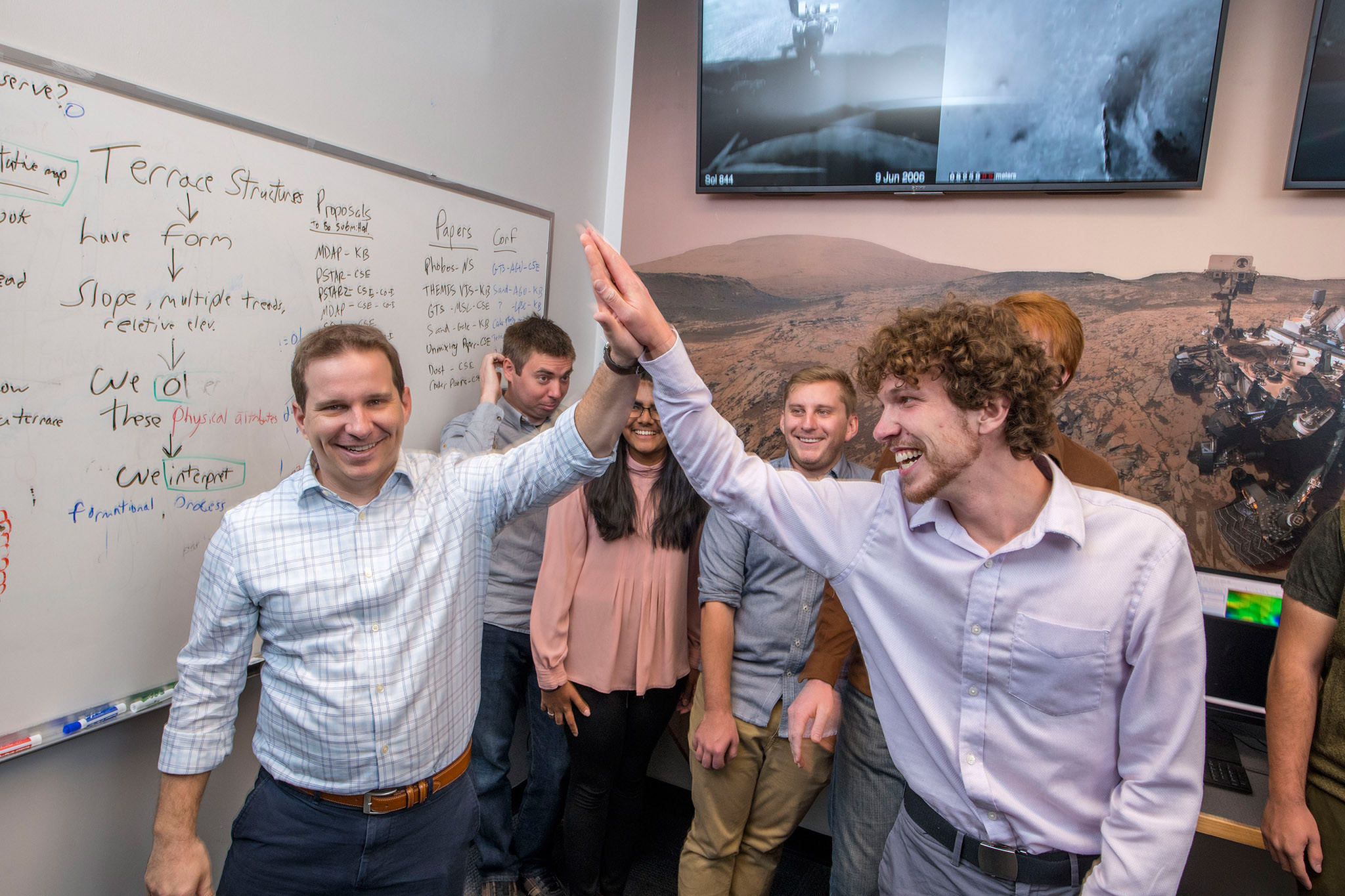 Students high fiving and smiling with a filled out whiteboard in the background.