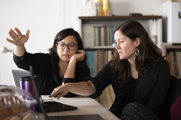 Two people, both wearing black clothing, sitting at a desk and looking at a laptop computer. One person is gesturing with their hand while the other person is resting their head on their hand. There are bookshelves in the background.