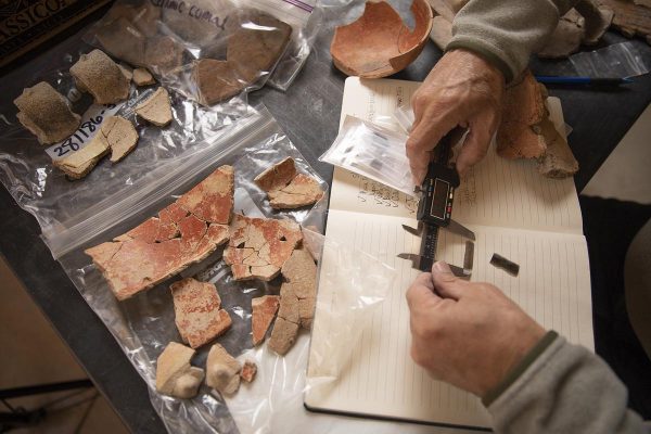 Hands holding a notebook and writing with a pen. Clear plastic bags containing various rocks and stone fragments. A dark-colored table or surface.