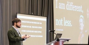 Daniel Wendler presenting in front of a CART captioning screen and next to a presentation screen that says I am different, not less.