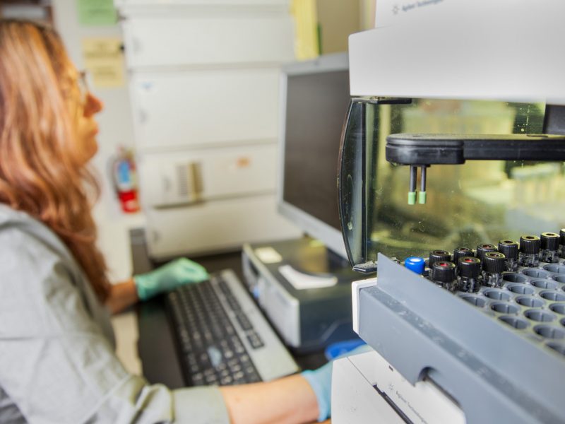 Researcher wearing gloves sitting at desktop aside large metal lab machine.