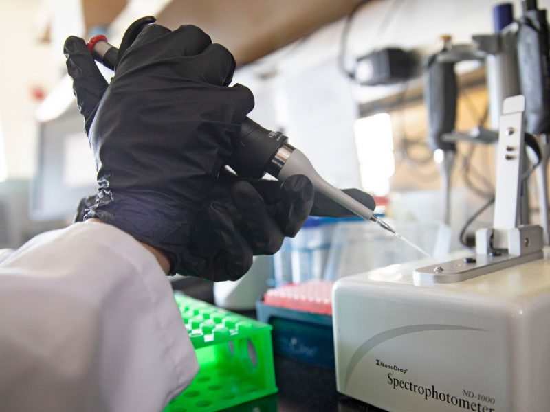 Gloved hands using syringe to put material into a machine in research lab.