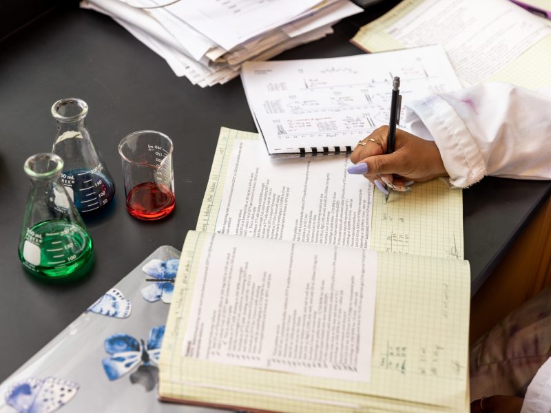 Hand writing in a book with glass containers of red, blue, and green liquid in a lab.