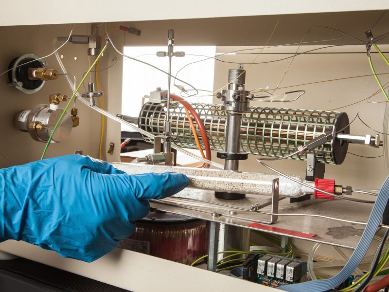 Gloved hand in lab using syringe alongside research equipment in lab.