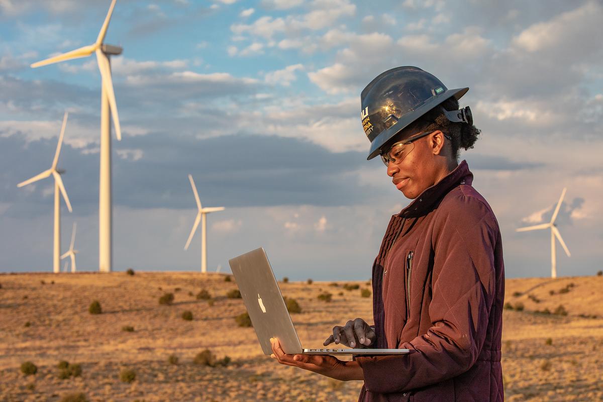 An N A U student, wearing a hardhat and looking at their computer, standing in a grassy plain with windmills behind them.