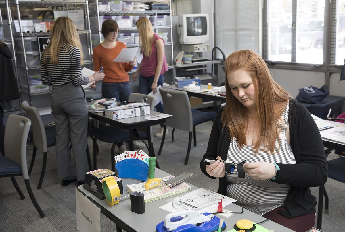 Three female students working on adaptive equipment on tables surrounded by various shelves, storage, and equipment visible in the background.