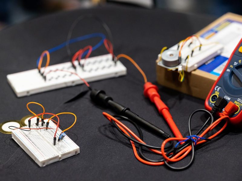 Assorted electronic components and tools, including a circuit board, wires, a multimeter, and other devices, arranged on a black surface.