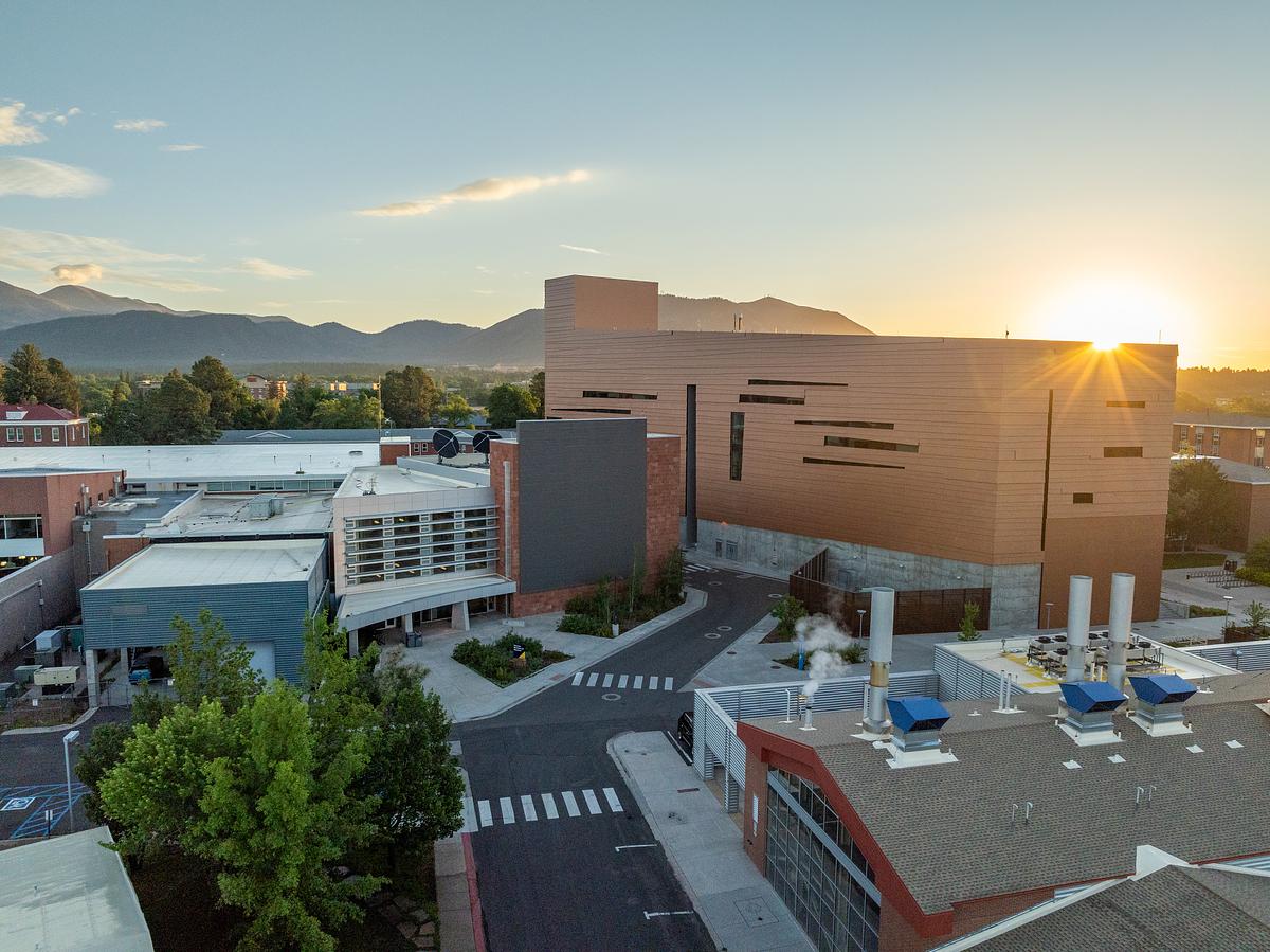 Aerial view of N A U Science and Health building and various buildings. It includes trees and a parking area at sunrise. The buildings feature a mix of materials and geometric designs, with some buildings having angled shapes and patterns. The surrounding mountains are visible in the background under a partly cloudy sky.