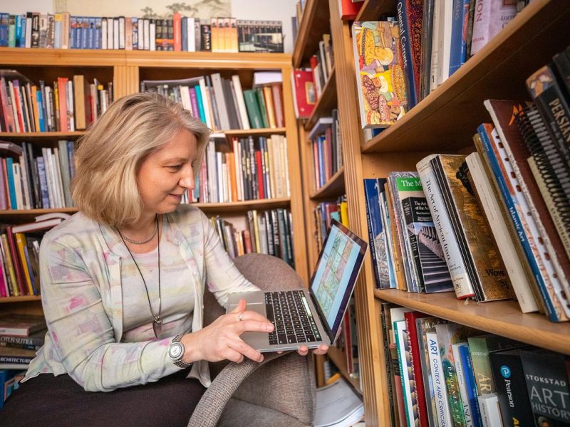 N A U researcher sitting in front of bookshelves, using a laptop computer. The bookshelves contain various books and items.