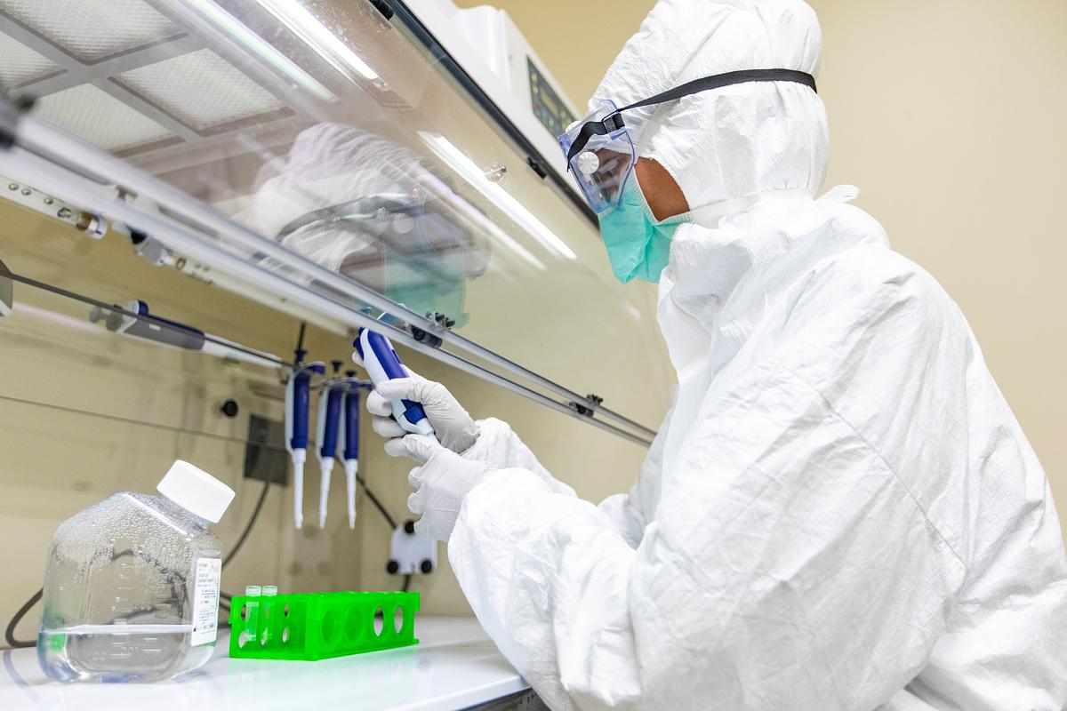Person wearing full protective suit, gloves, and mask working in a laboratory with various equipment and materials.