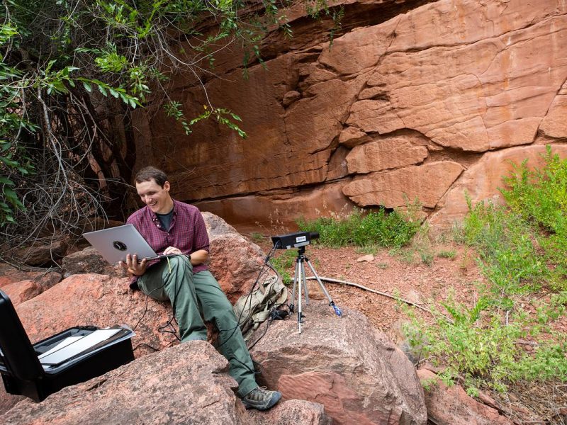 Person sitting on a rock in a natural setting, using a laptop computer on a tripod. Red rock formations and vegetation in the background.