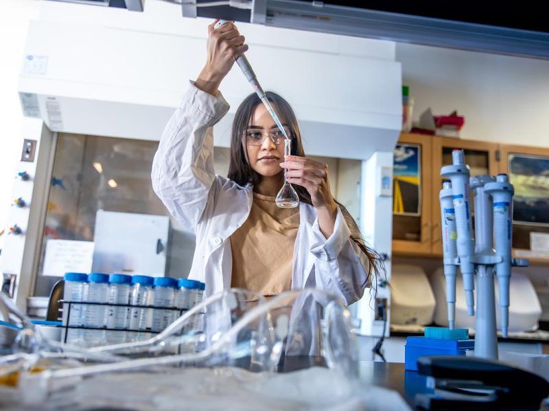Individual in a laboratory setting, wearing a white coat and safety goggles, examining a test tube and conducting scientific experiments.