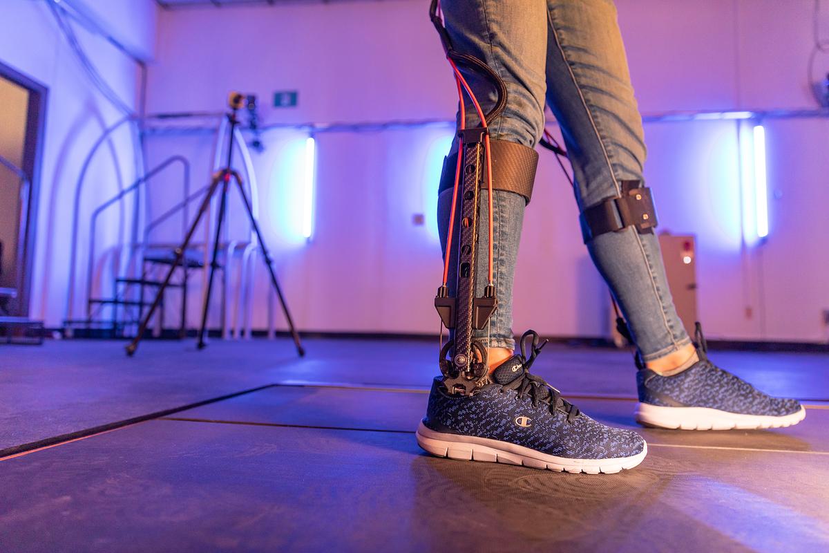 Person' wearing an exoskeleton in Zach Lerner's lab. They are wearing casual clothing and sneakers, standing on a tiled floor in a room with neon lighting.