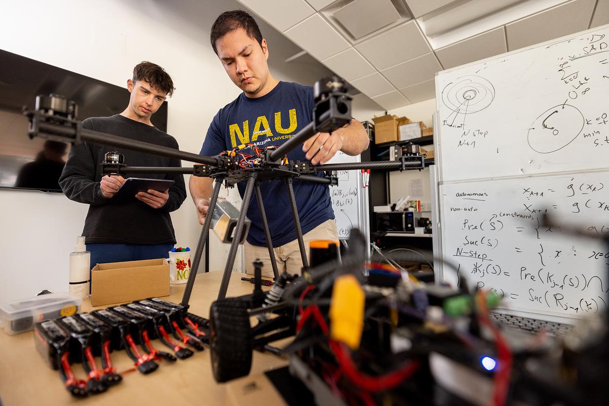 Two students working on a project involving a drones, tools, and a whiteboard with equations and notes. One individual is holding a tablet and the other is manipulating a piece of equipment on a table. The individuals are wearing casual clothing, including a sweatshirt with the text "NAU".