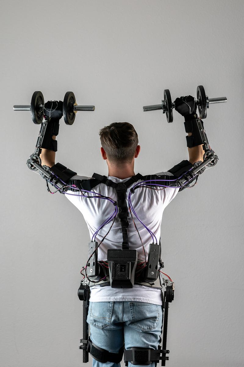 A person wearing a white shirt, jeans, and a an exoskeleton weight-lifting harness. The person is holding weighted dumbbells above their head, with their arms extended upwards.