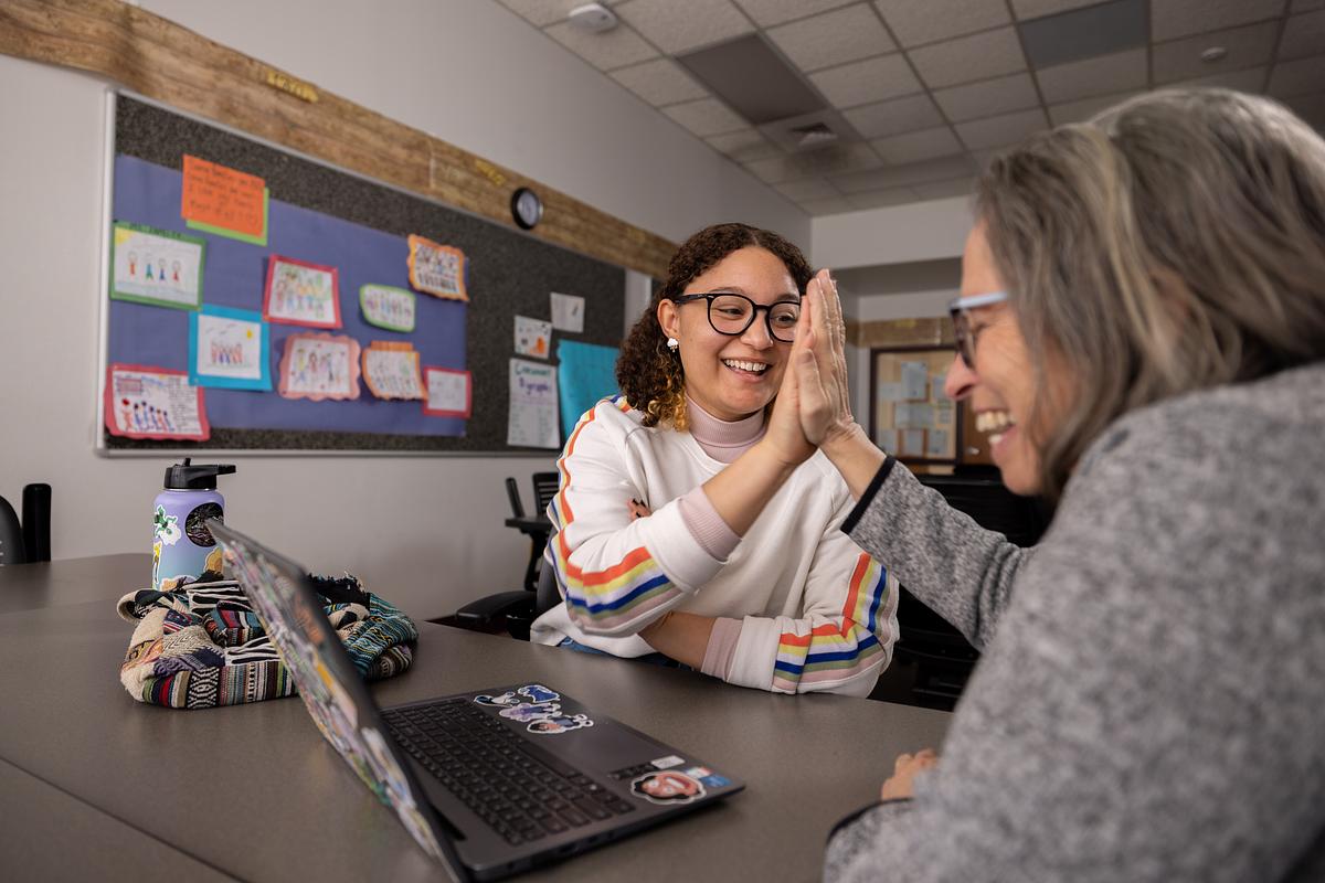 Two people high-fiving each other. A computer sits on the table and colorful pieces of paper may be seen on a board in the background.