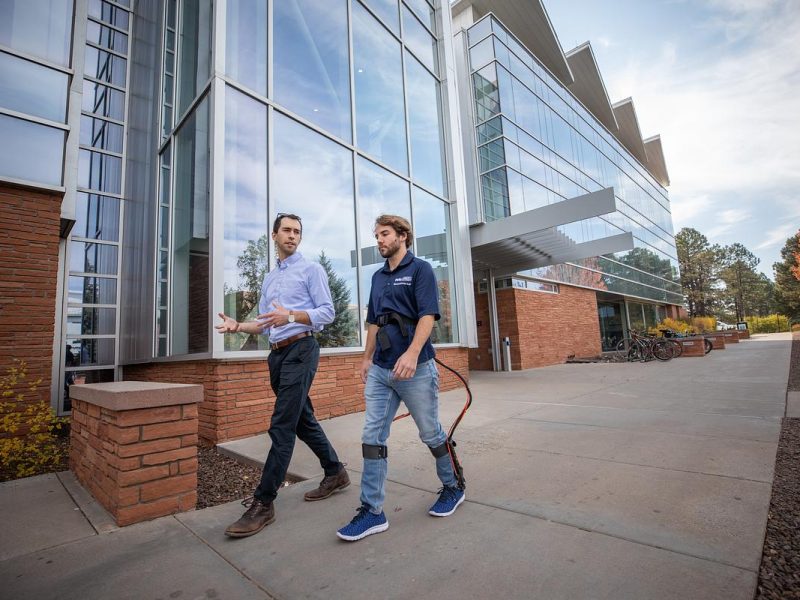 Zach Lerner walking with a student researcher in front of the engineering building. The student is wearing an exoskeleton.