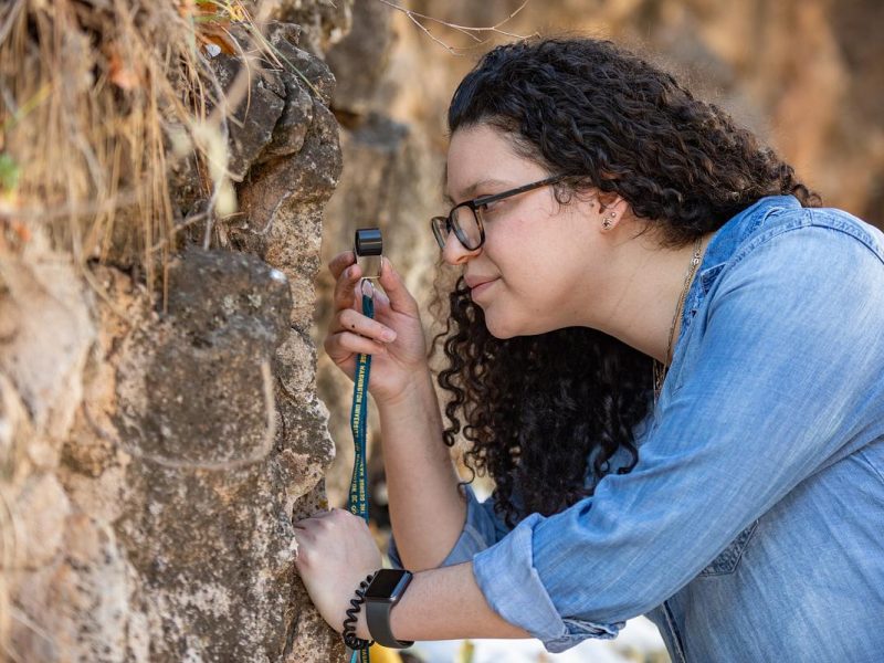Graduate research student with curly dark hair, wearing glasses and a denim jacket, examining a rock surface with a tool in the field.