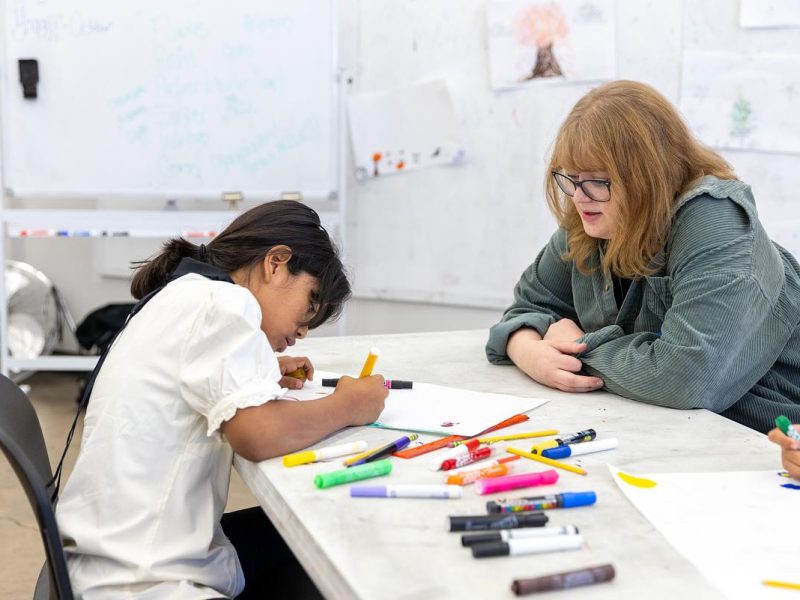 Two individuals, one sitting at a table using colored markers and the other observing, in a room with art supplies and artwork on the walls.