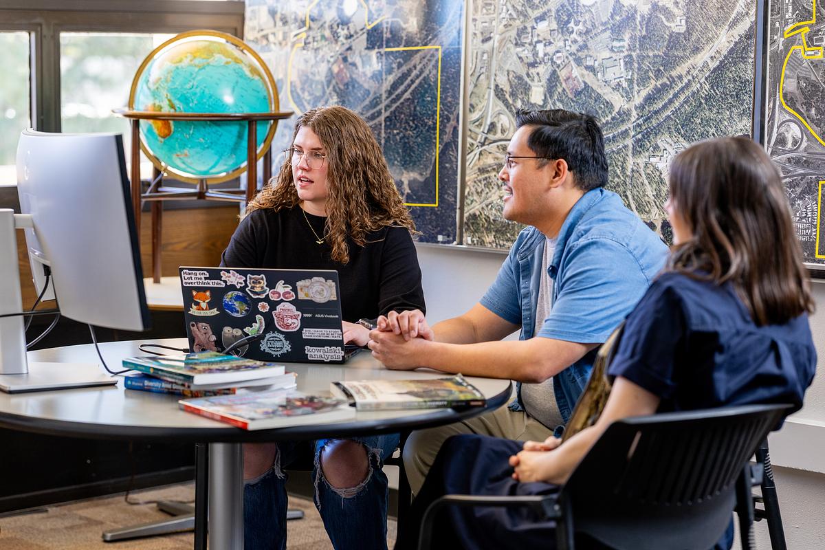 Three research students sitting at a table in an office or classroom setting engaged in discussion. A laptop computer, stickers, and papers are visible on the table.