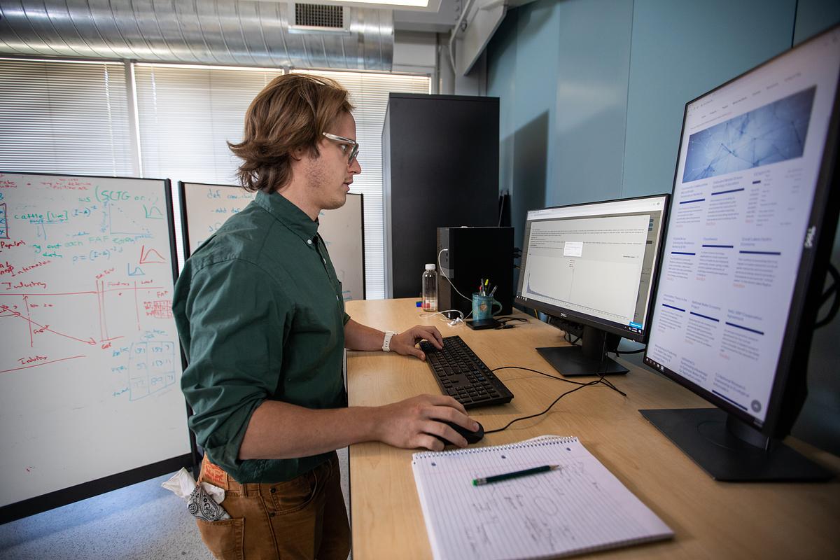 Student in a green shirt working at a computer work stations in a research lab.
