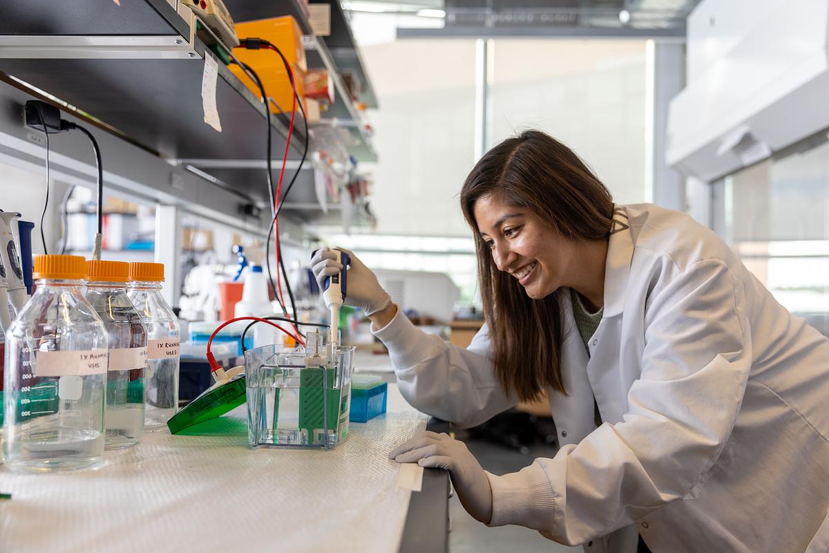 Student in white lab coat working at lab bench with scientific equipment and supplies.