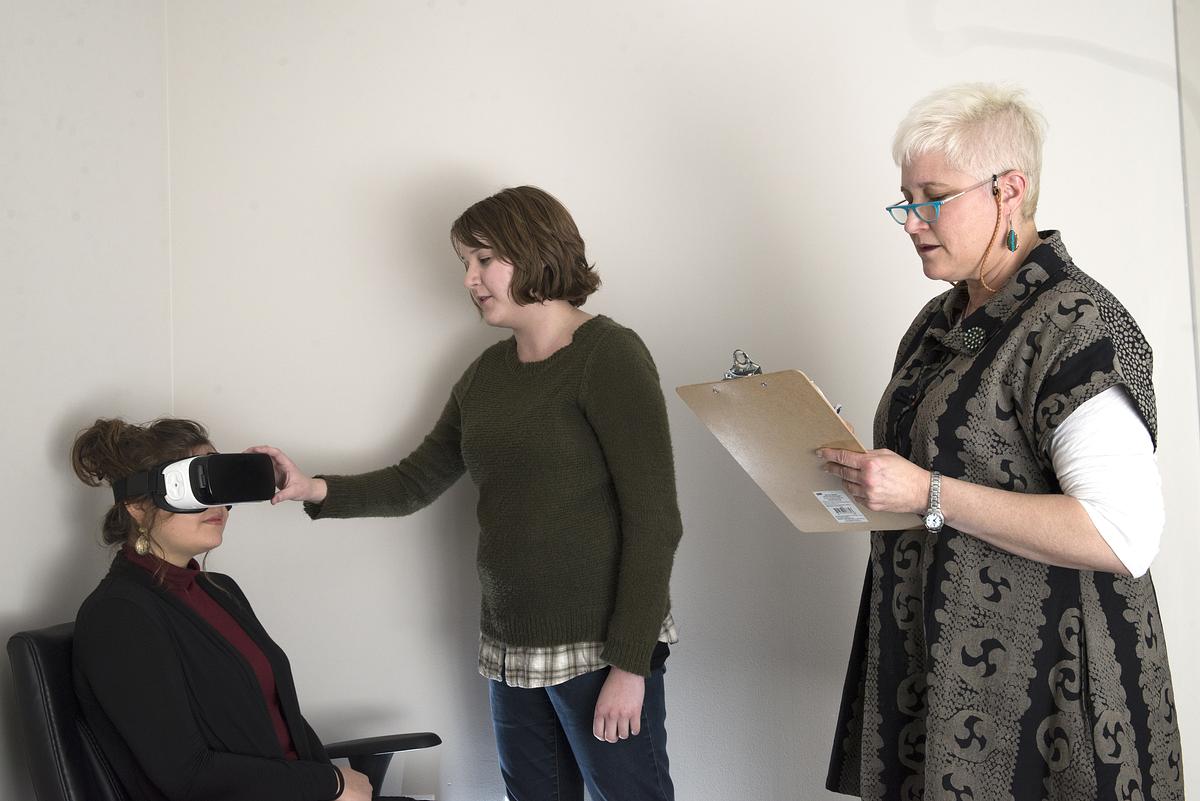 Researchers working with a student wearing VR glasses that is sitting at a desk.