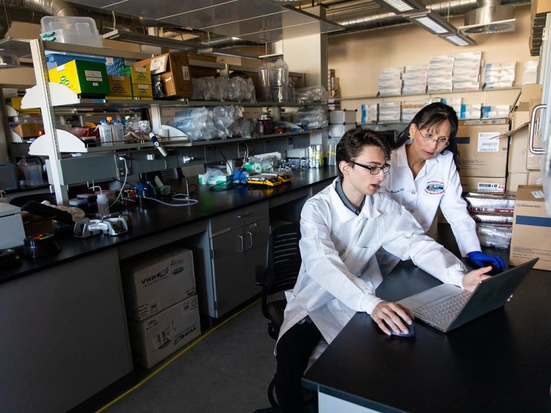 Two individuals in white lab coats, sitting on a desk in a laboratory setting, examining a laptop computer.