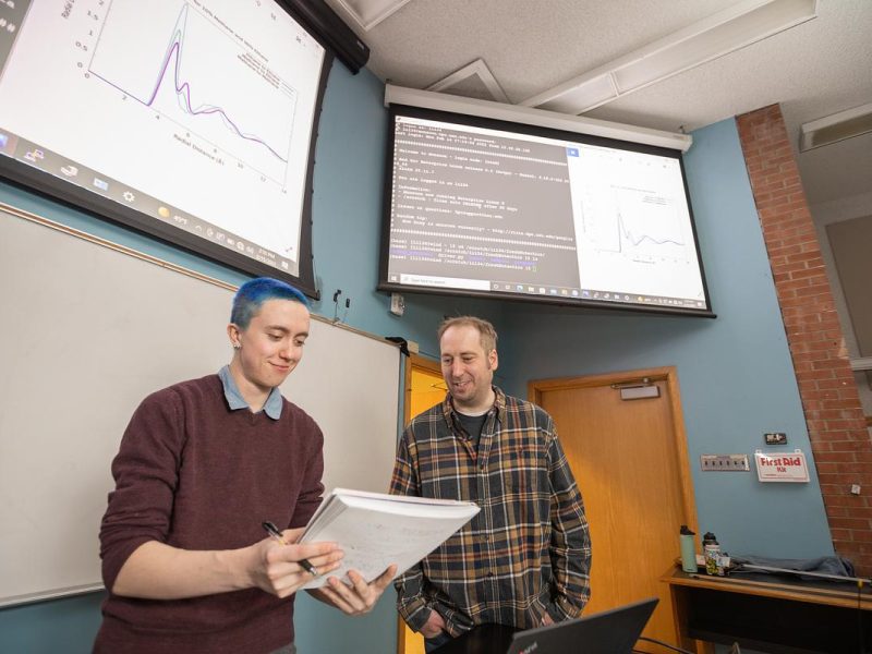 Student standing with faculty mentor, one with short hair and one with longer hair, standing in a room with a large display showing graphs and text for the undergraduate symposium.