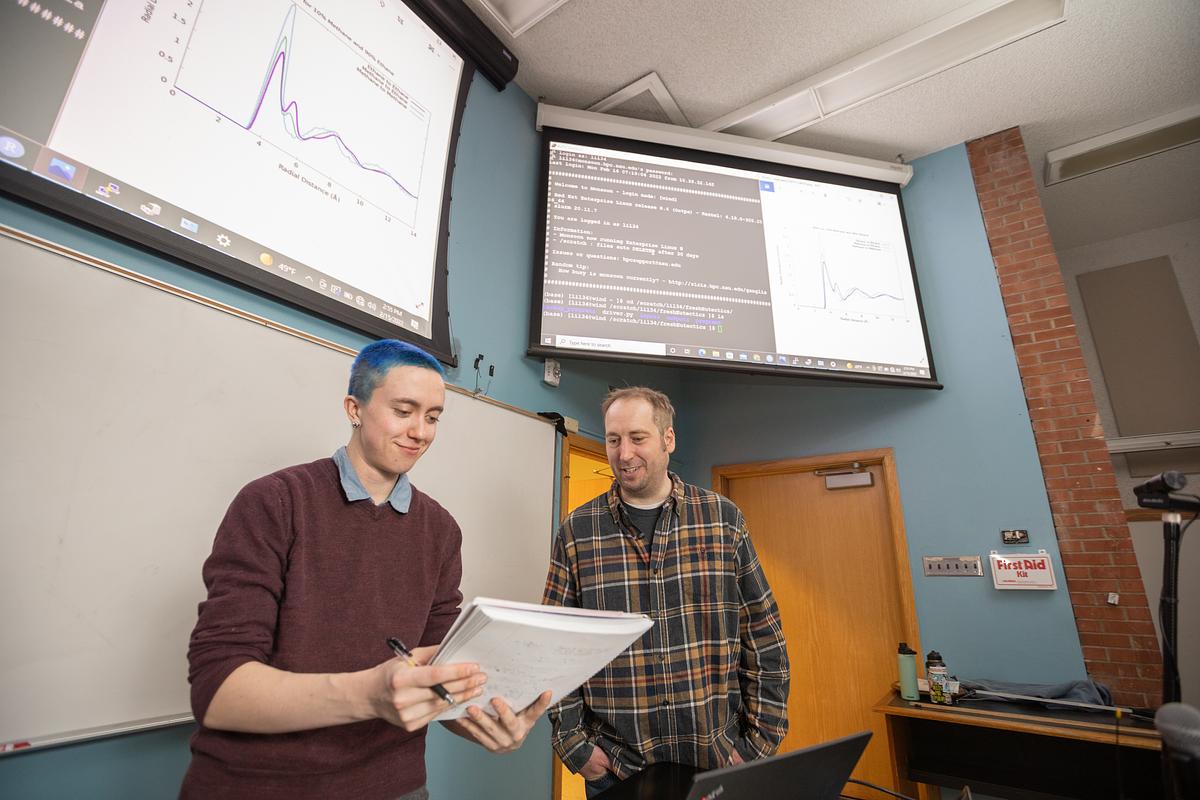 Student standing with faculty mentor, one with short hair and one with longer hair, standing in a room with a large display showing graphs and text for the undergraduate symposium.