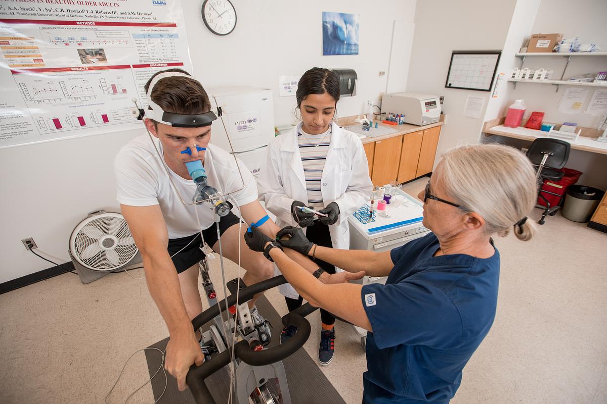 Two people wearing lab uniforms and protective equipment, assisting an adult in using a piece of exercise equipment in a medical facility. A chart and shelving units can be seen in the background.